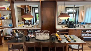 Un'ampia sala da pranzo con un lungo tavolo apparecchiato per un pasto, circondato da sedie di legno. Sul tavolo è posizionata una grande ciotola di metallo piena di bottiglie e bicchieri, e si nota anche un vassoio con diverse ciotole di cibo.