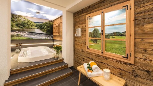 Une salle de spa dotée d’une grande baignoire, de murs en bois, d’une fenêtre donnant sur un paysage verdoyant et d’une petite table avec des serviettes et une bougie.
