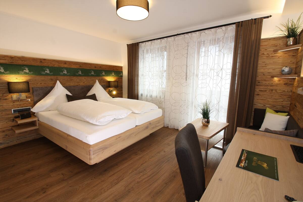 Ein Hotelzimmer mit einem hölzernen Bett, einem Schreibtisch mit Stuhl, einem kleinen Tisch und einer Topfpflanze.