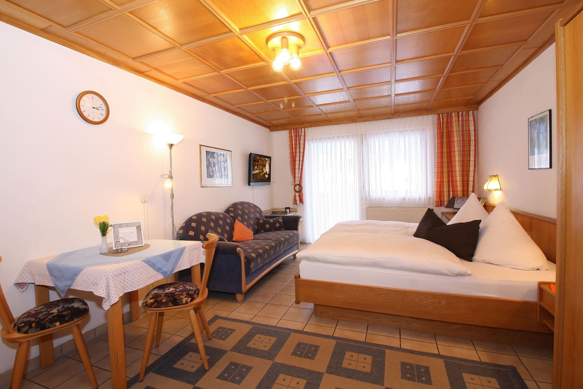 Ein Zimmer mit Bett, Schreibtisch mit Stuhl, Sofa, einer Uhr und einem Fenster mit Vorhängen.