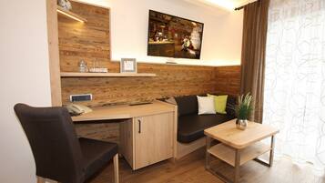 A compact room with a wooden accent wall, a mounted TV, a desk with a chair, a sofa, and a small table with a plant.