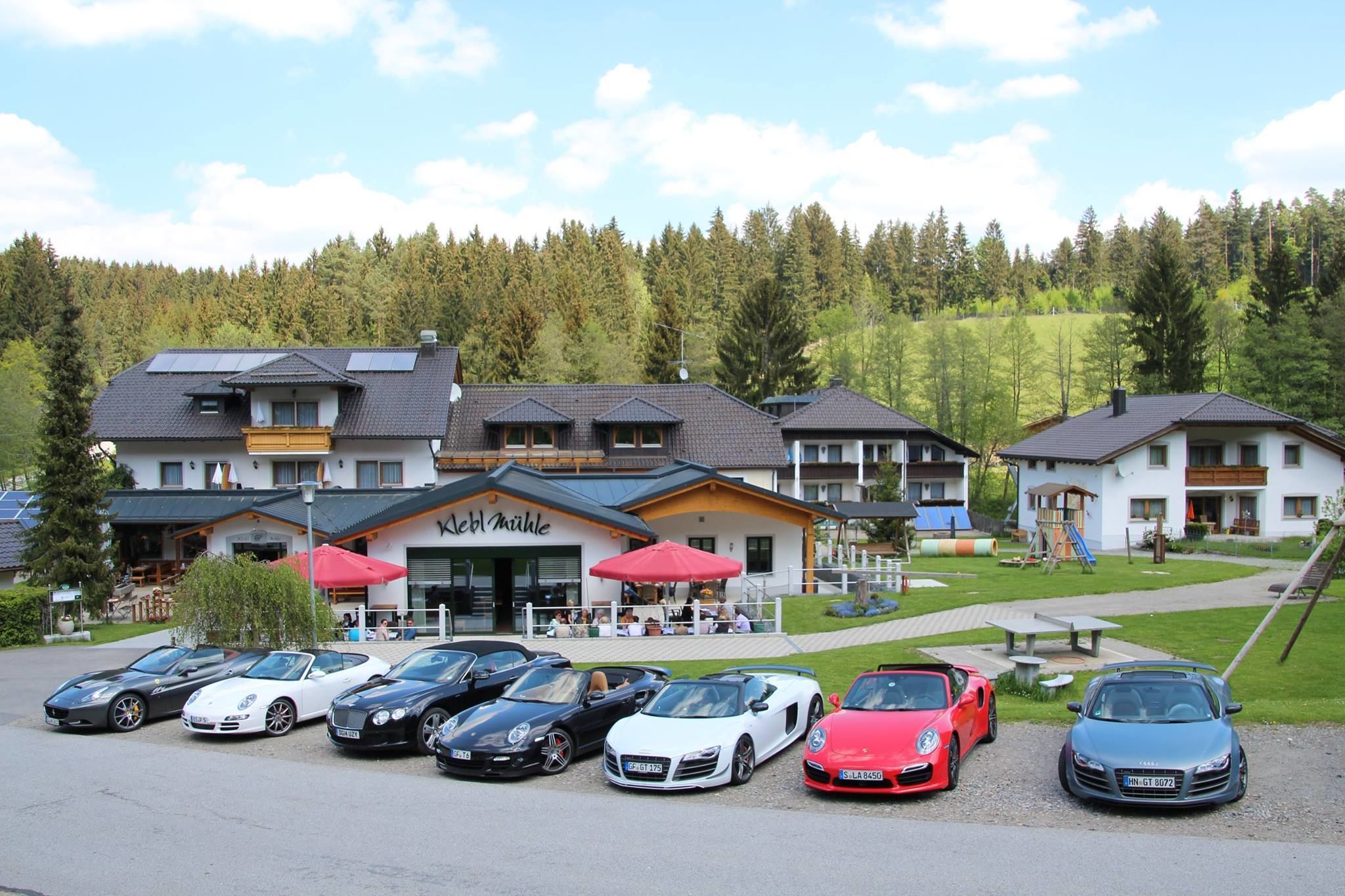Et hotel med flere bygninger, en parkeringsplads med flere luksusbiler og en skovklædt bakke i baggrunden.