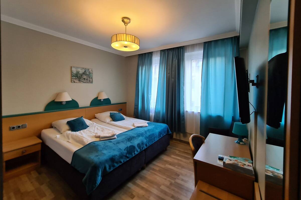 A hotel room with a bed, two bedside tables, a desk, a chair, a television, and a window with curtains.
