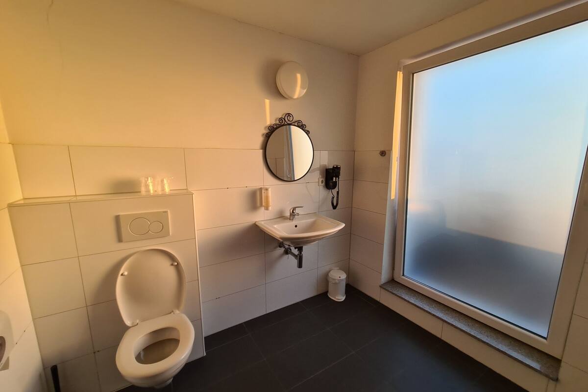 A bathroom with a toilet, sink, and mirror. There is a window with frosted glass and a wall-mounted phone.
