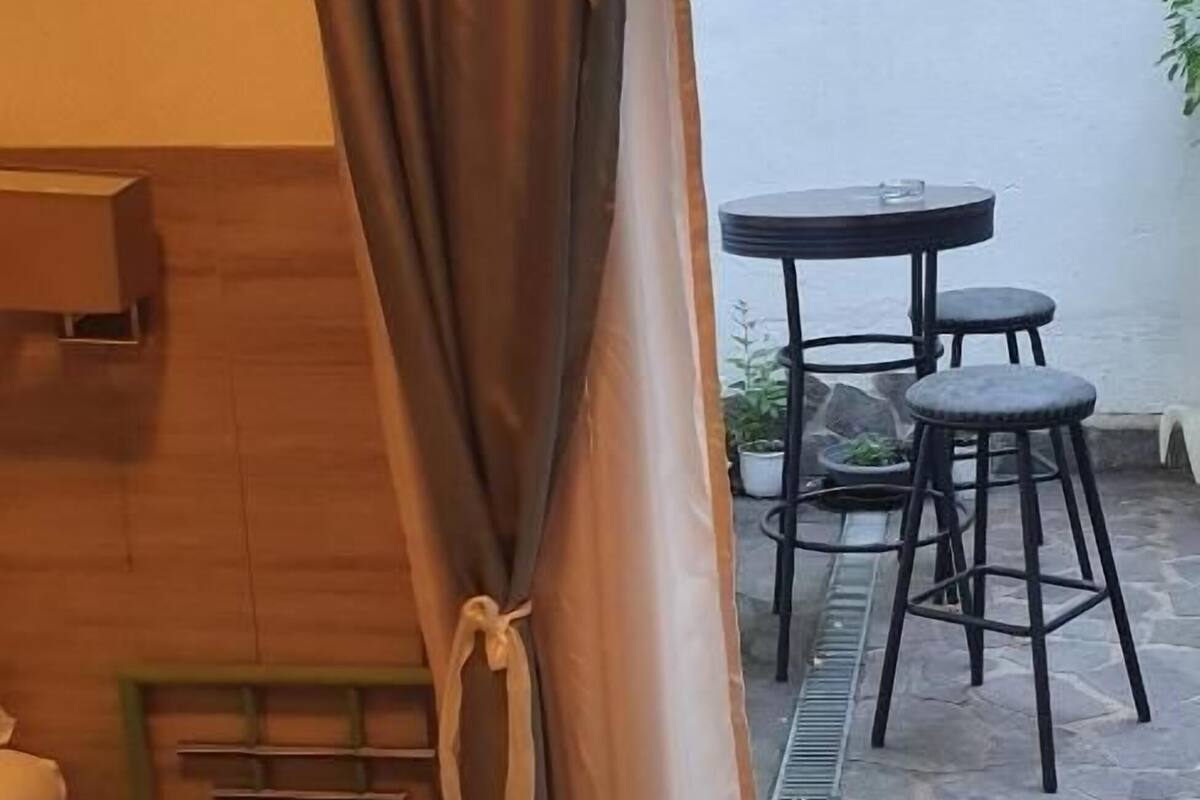 A room with a bed, a small table, a green stool, a curtain, and a view of an outdoor patio with a table and chairs.