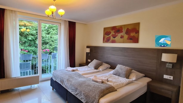 A hotel room with a large bed, two bedside tables, a radiator, and a view of a balcony with potted plants. - Cara Vita Hotel (Cologne)