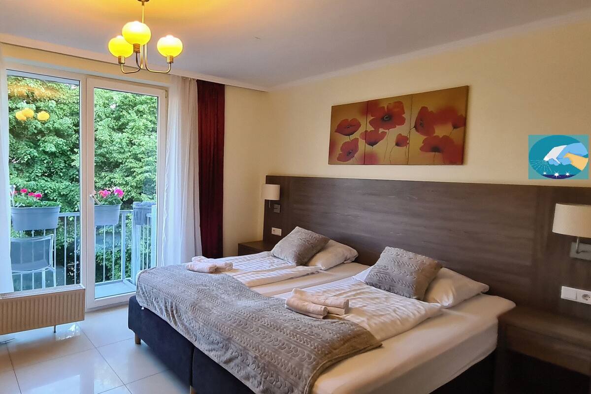A hotel room with a large bed, two bedside tables, a radiator, and a view of a balcony with potted plants.