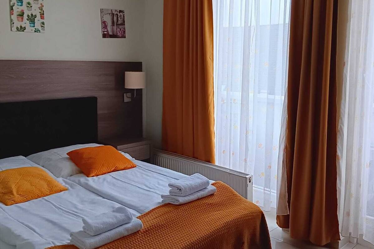 A hotel room with a bed, orange curtains, a radiator, and a window with sheer curtains.