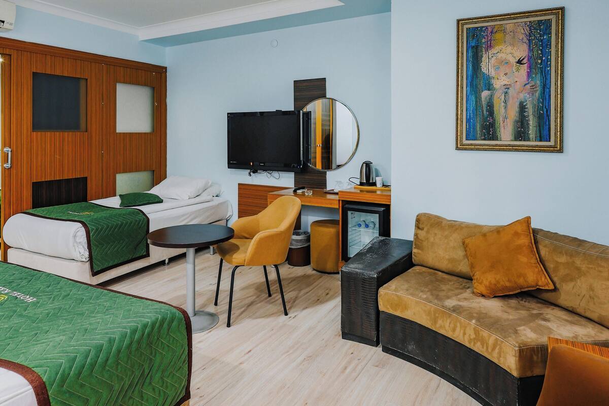 A hotel room with two beds, a desk with a computer, a chair, a sofa, a painting on the wall, and a ceiling light.
