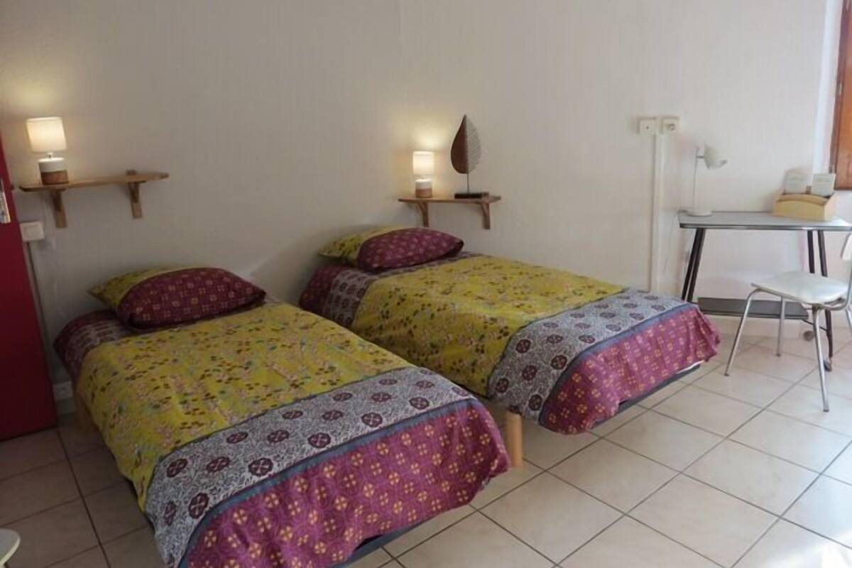 A room with two beds, each with patterned bedding and pillows, a desk with a chair, and wall-mounted lamps.