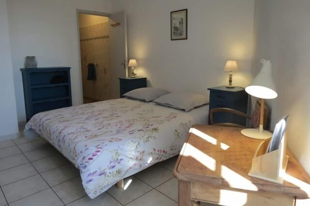 A bedroom with a bed, two bedside tables with lamps, a wooden chair, a small table with a lamp, and a framed picture on the wall.
