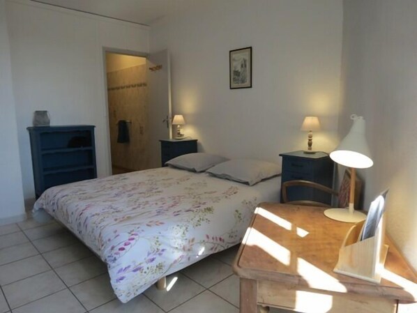 Une chambre à coucher avec un lit, deux tables de chevet avec des lampes, une chaise en bois, une petite table avec une lampe et un tableau encadré au mur.