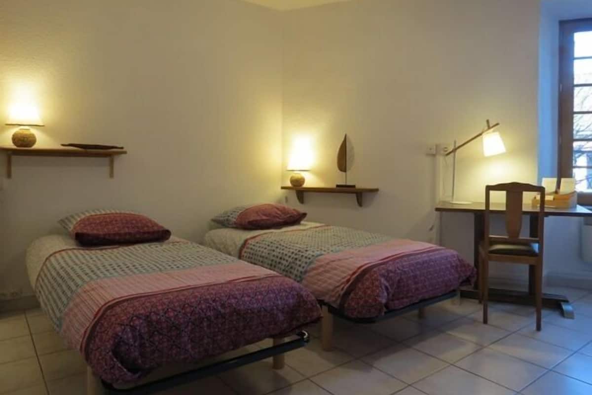 A small, well-lit room with two beds, each with a patterned bedspread and pillows, a desk with a chair, and wall-mounted shelves with lamps.