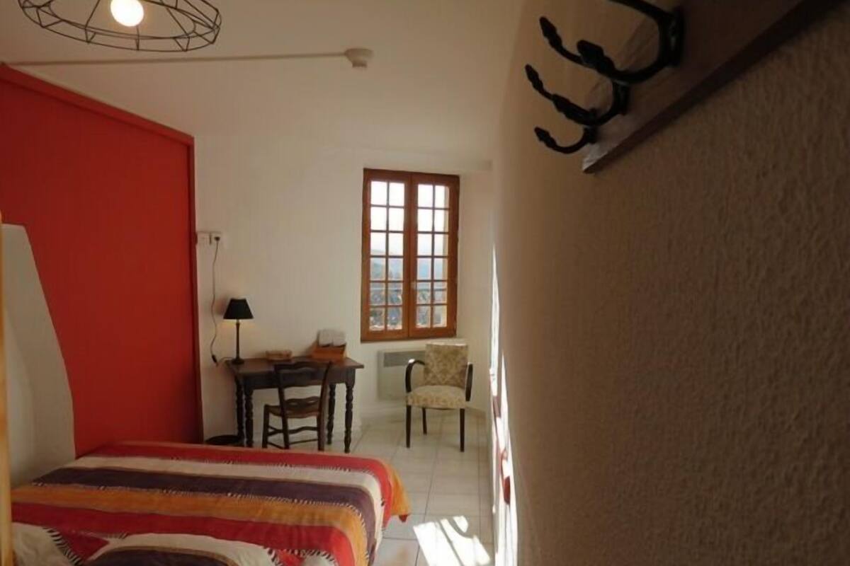 A bedroom with a bed, a desk, and a chair. There is a window with a view of the outside.