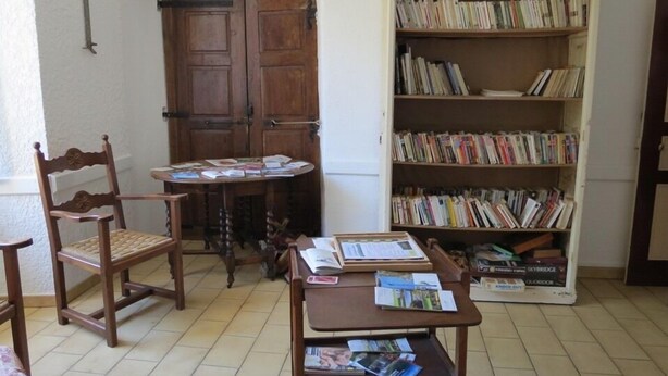 A room with a wooden chair, a small round table, and a bookshelf filled with books. - Cafe Restaurant Meuble de Tourisme (Saint-Germain-de-Calberte)
