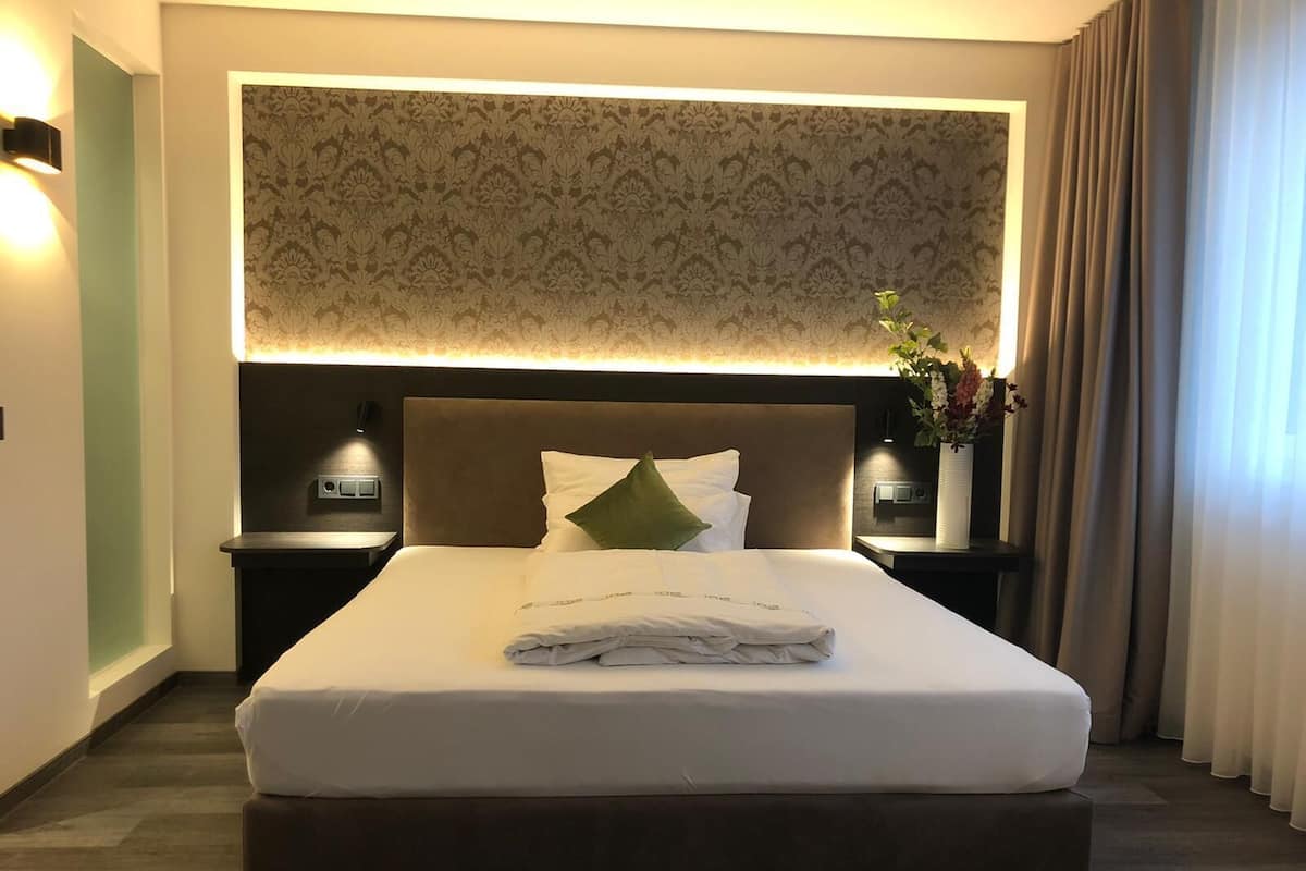 A hotel room with a large bed, a headboard with a patterned wallpaper, bedside tables with lamps, and a vase with flowers on the right side.