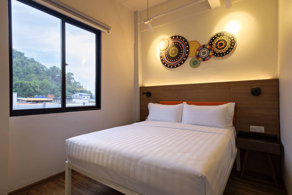 A hotel room with a large bed, a nightstand, a wall-mounted light fixture, and a window with a view of buildings and greenery.
