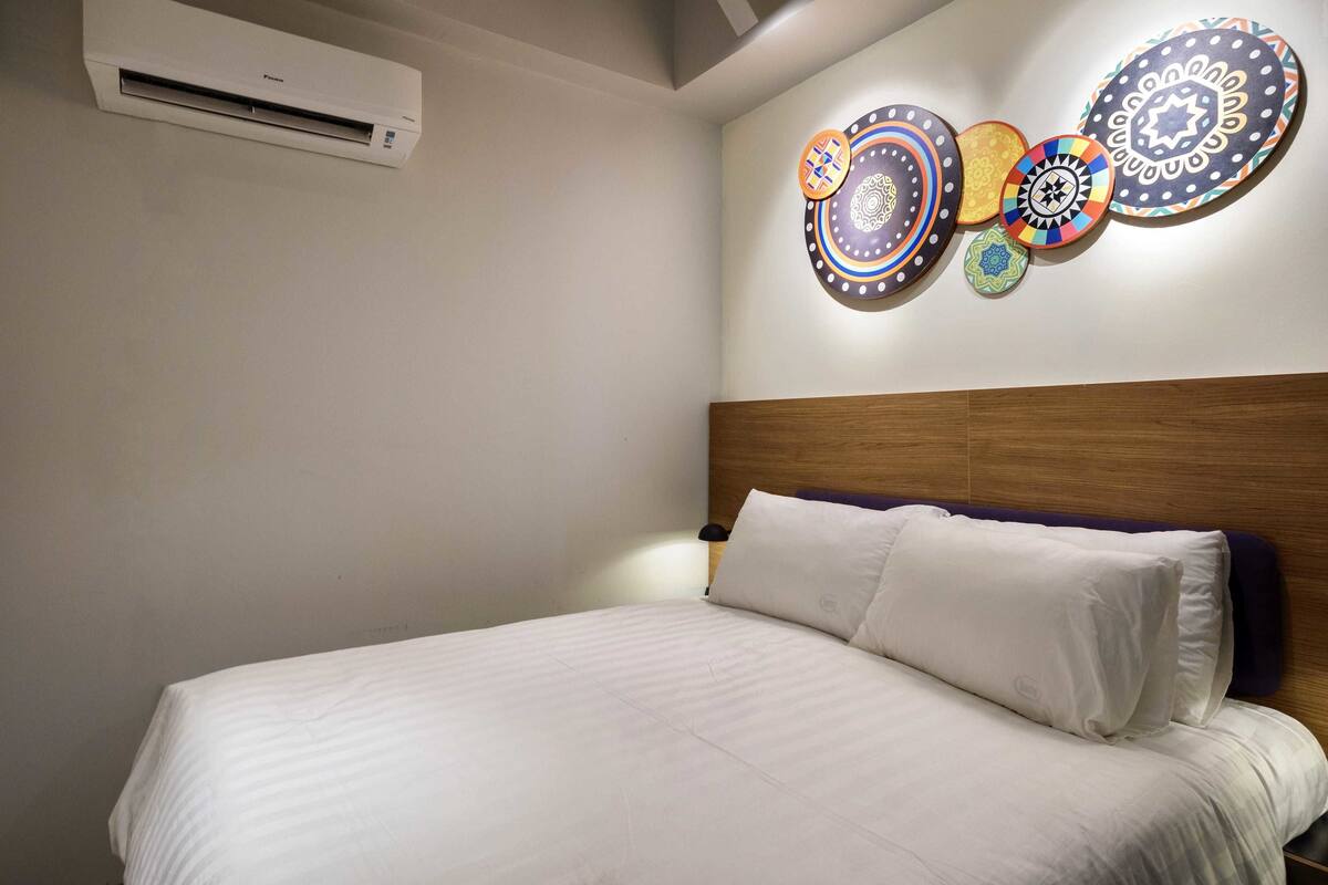 A bedroom with a bed, white bedding, a wooden headboard, a wall-mounted air conditioner, and a decorative wall with circular patterns.