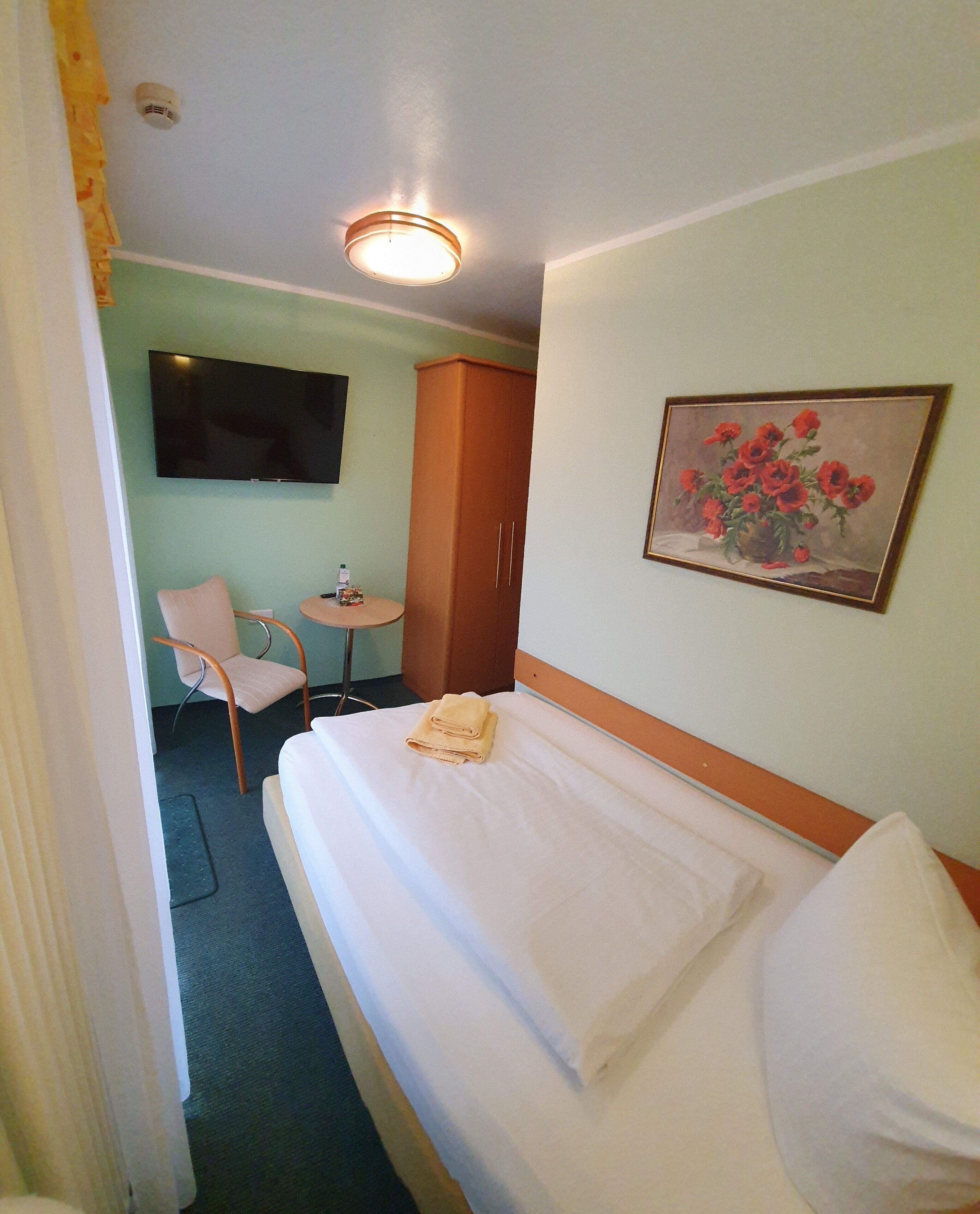Quarto de hotel com cama, uma cadeira, mesa pequena, guarda-roupa, um quadro e televisão na parede.