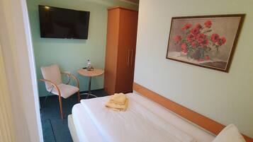 Quarto de hotel com cama, uma cadeira, mesa pequena, guarda-roupa, um quadro e televisão na parede.