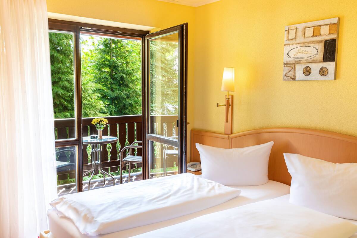 Ein Hotelzimmer mit Bett, Nachttisch, Lampe, Fenster mit Blick auf Grünflächen und einem kleinen Tisch mit Stühlen draußen.