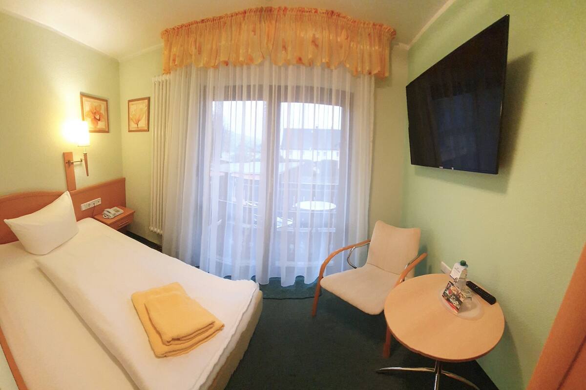 Ein Hotelzimmer mit Bett, Sessel, kleinen Tisch, Fernseher und Fenster mit Vorhängen.
