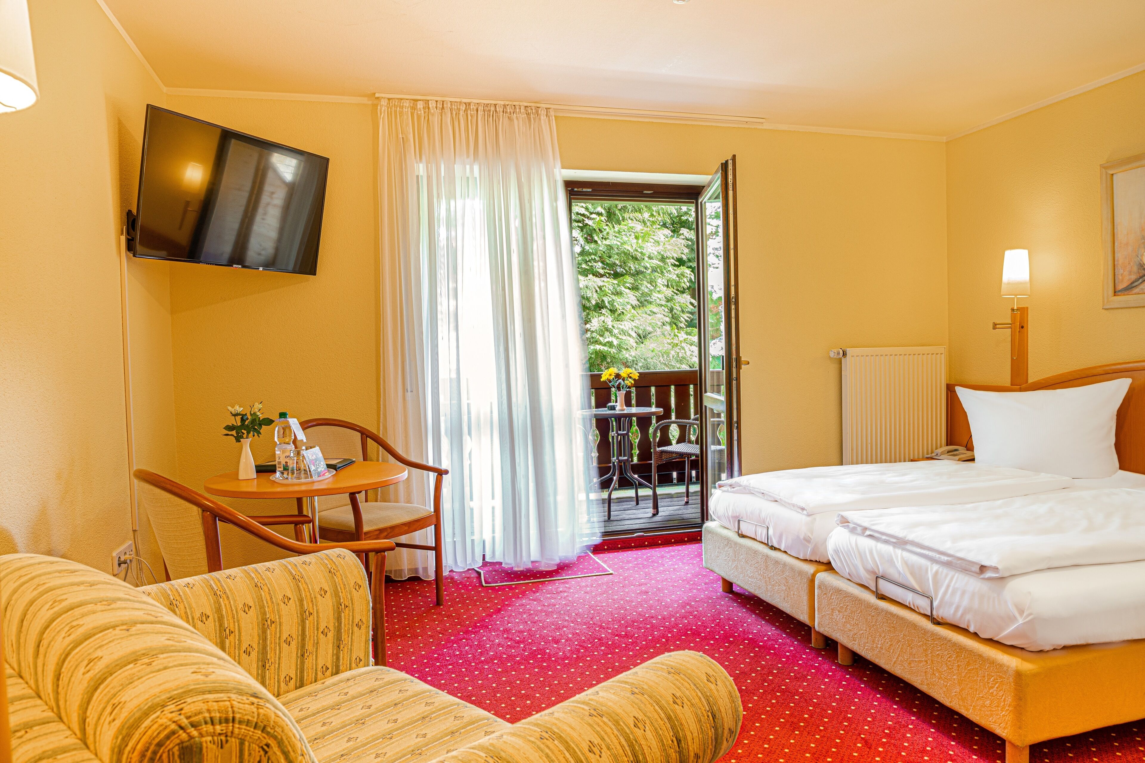 Ett hotellrum med en TV som är monterad på väggen, ett skrivbord med en stol, två sängar, en röd matta och utsikt över grönområden utanför.