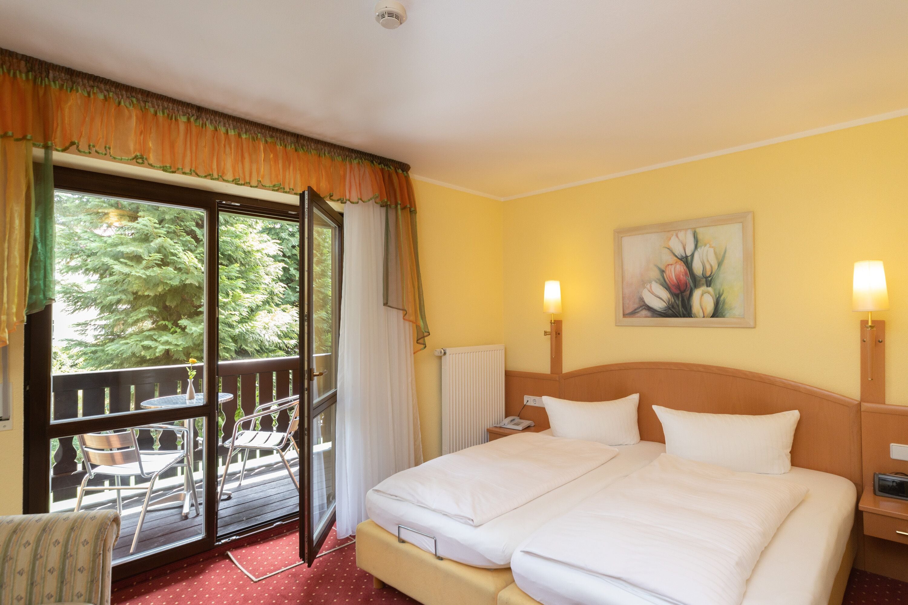A hotel room with a bed, two bedside lamps, a painting on the wall, a window with curtains, and a view of trees.