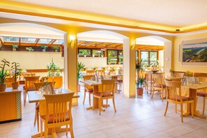 A spacious dining area with wooden tables and chairs, potted plants, and large windows allowing natural light. - Hotel Elbpromenade (Bad Schandau)