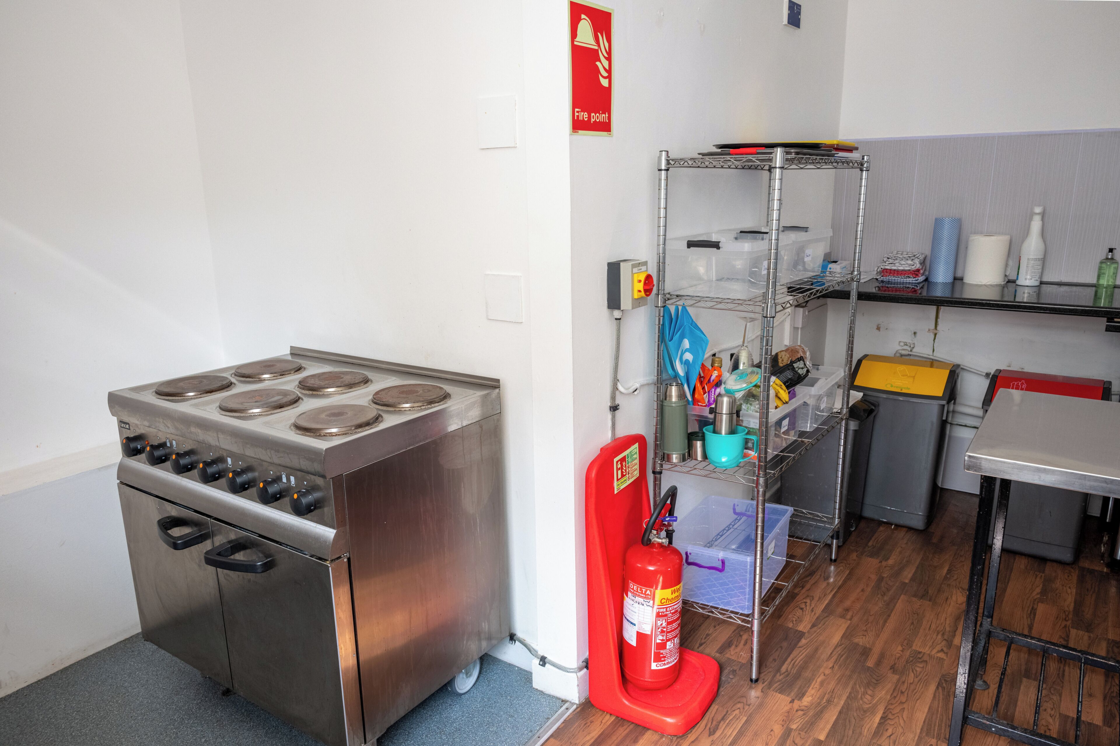 A kitchen with a stainless steel stove, a fire extinguisher, and a metal shelf with various items.