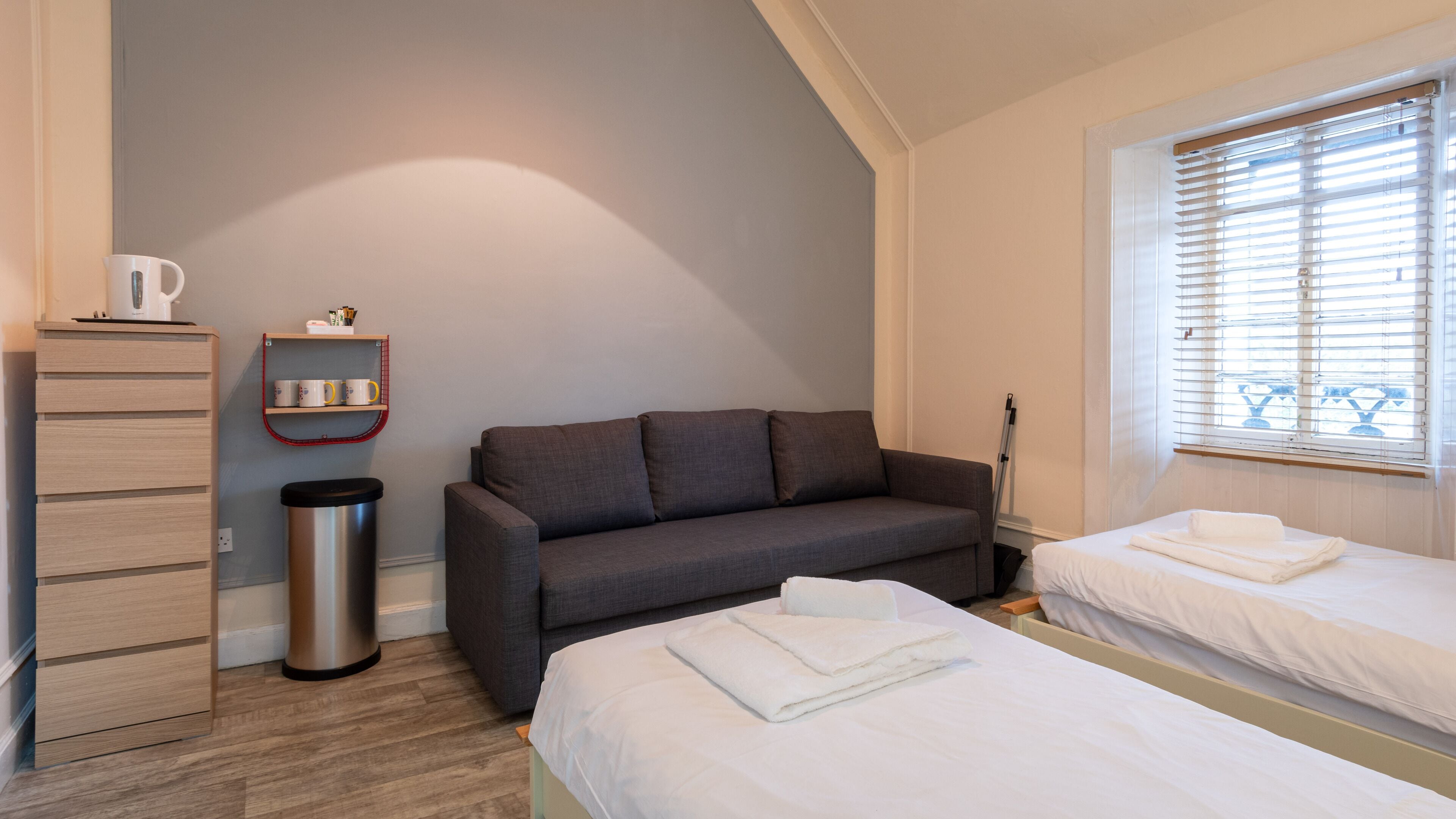 A room with a grey sofa, a small table with a kettle, a trash bin, and a window with blinds.