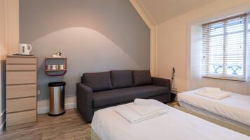 A room with a grey sofa, a small table with a kettle, a trash bin, and a window with blinds.
