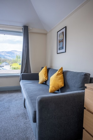 A grey sofa with two yellow pillows, a framed picture on the wall, and a window with a view of mountains. - Callander Hostel (Callander)