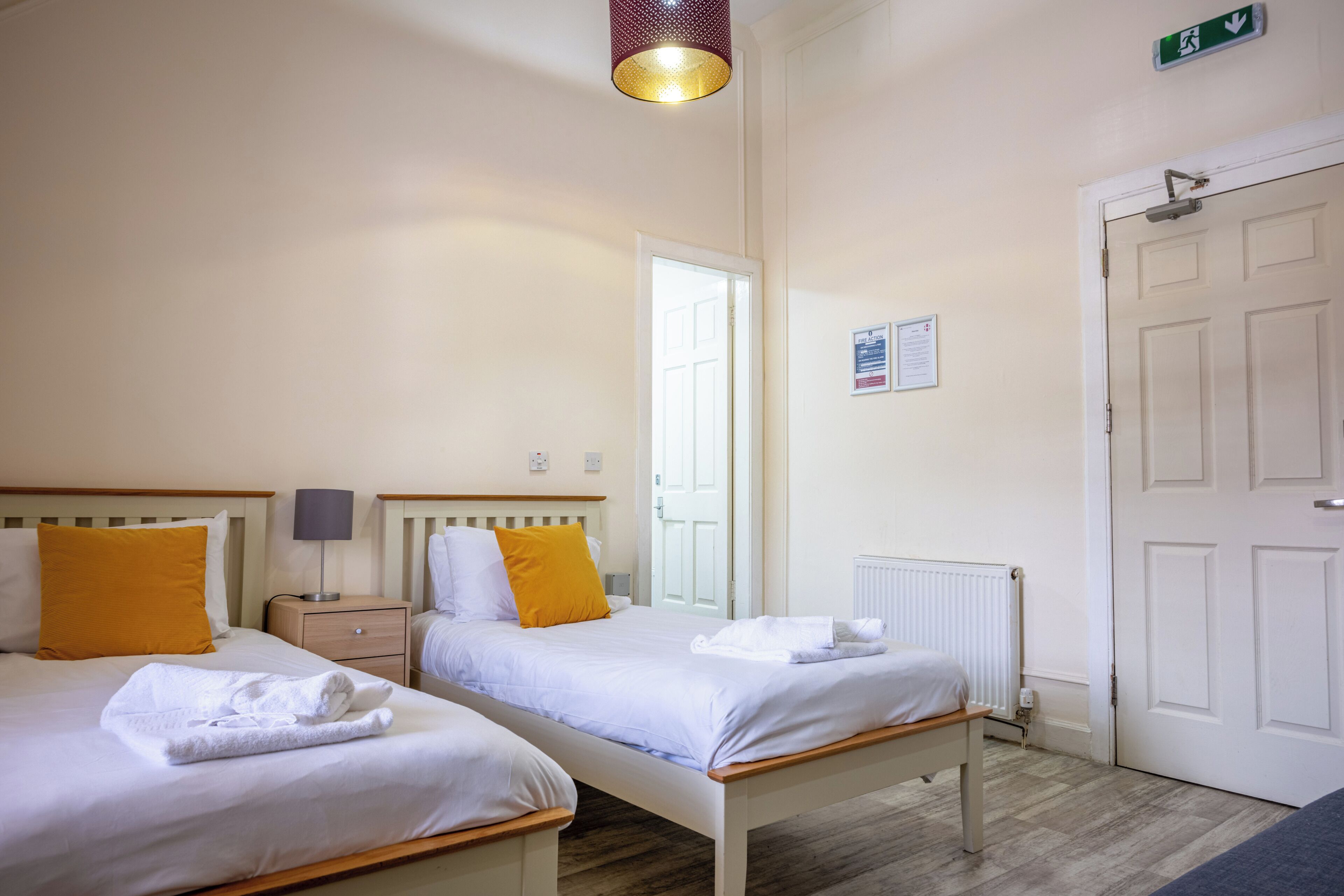 Habitación de hotel con dos camas, una mesita de noche con lámpara, un radiador y una puerta con letrero de salida de emergencia.