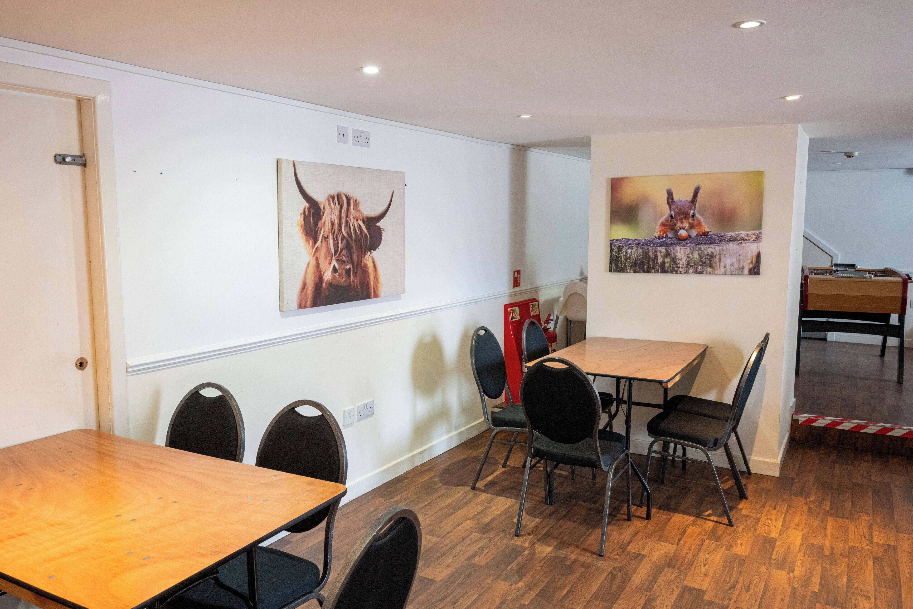 Un comedor con mesas y sillas de madera, dos cuadros en la pared con motivos de animales y un extintor.