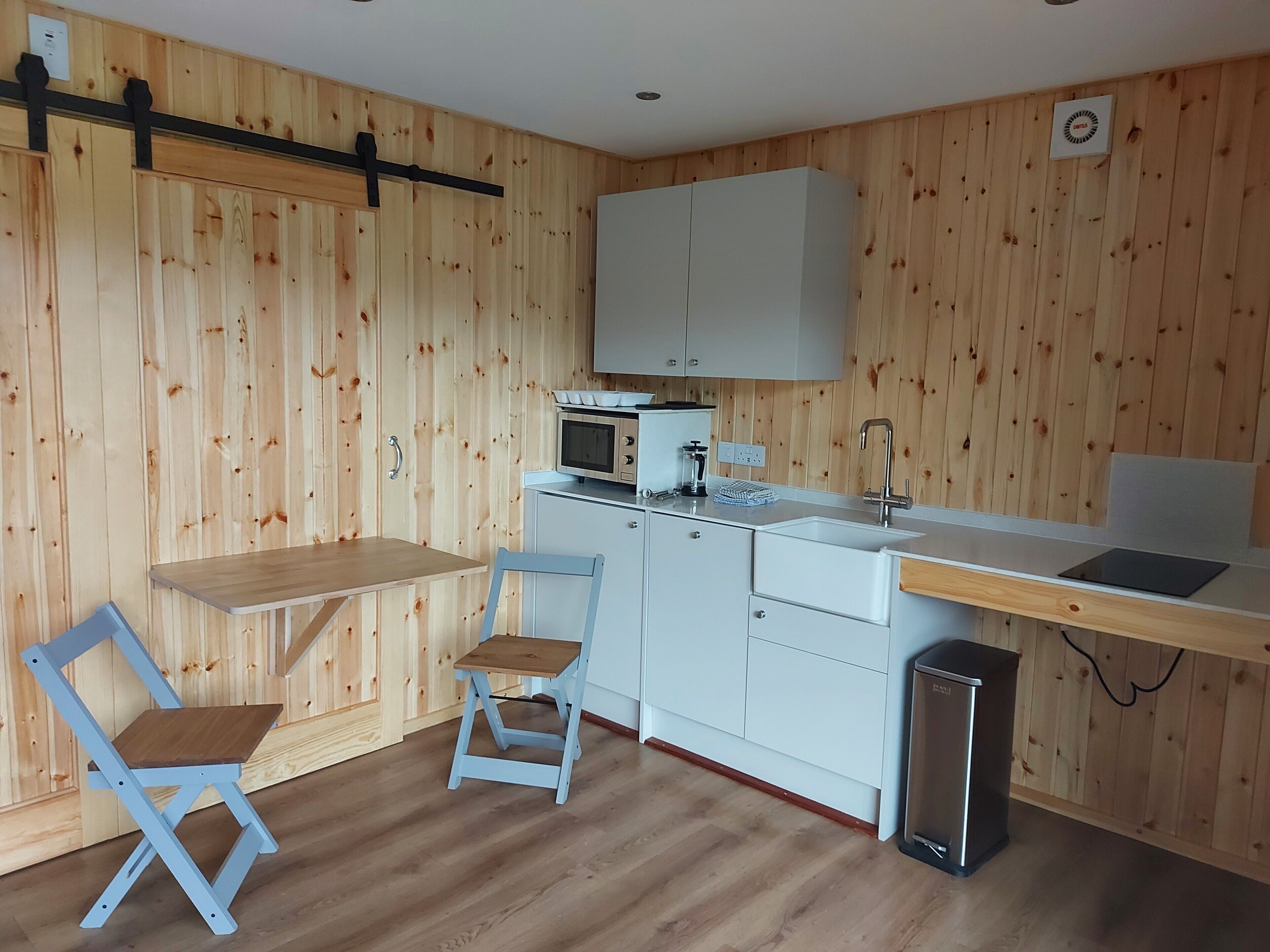 A compact kitchen with wooden panel walls, a white countertop, a sink, a microwave, and a trash bin.