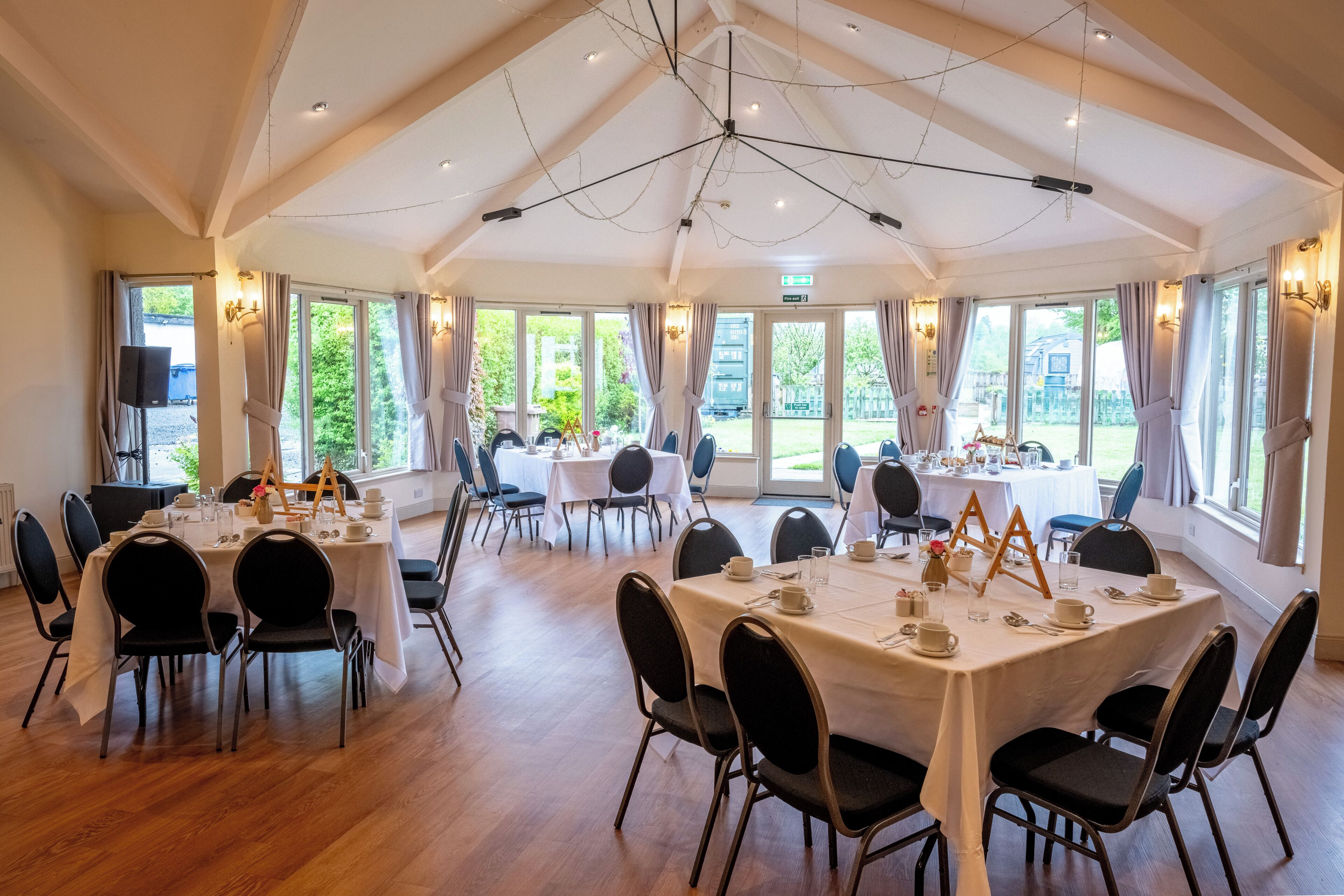 A spacious dining area with multiple tables and chairs, large windows with curtains, and a high ceiling with exposed beams.
