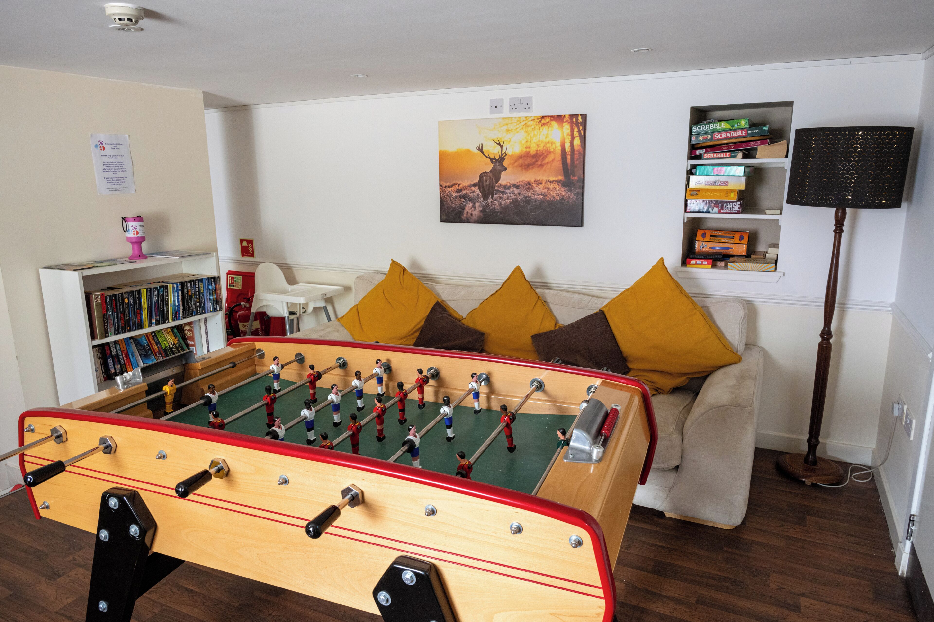 A room with a foosball table, a sofa with cushions, a bookshelf, and a painting on the wall.