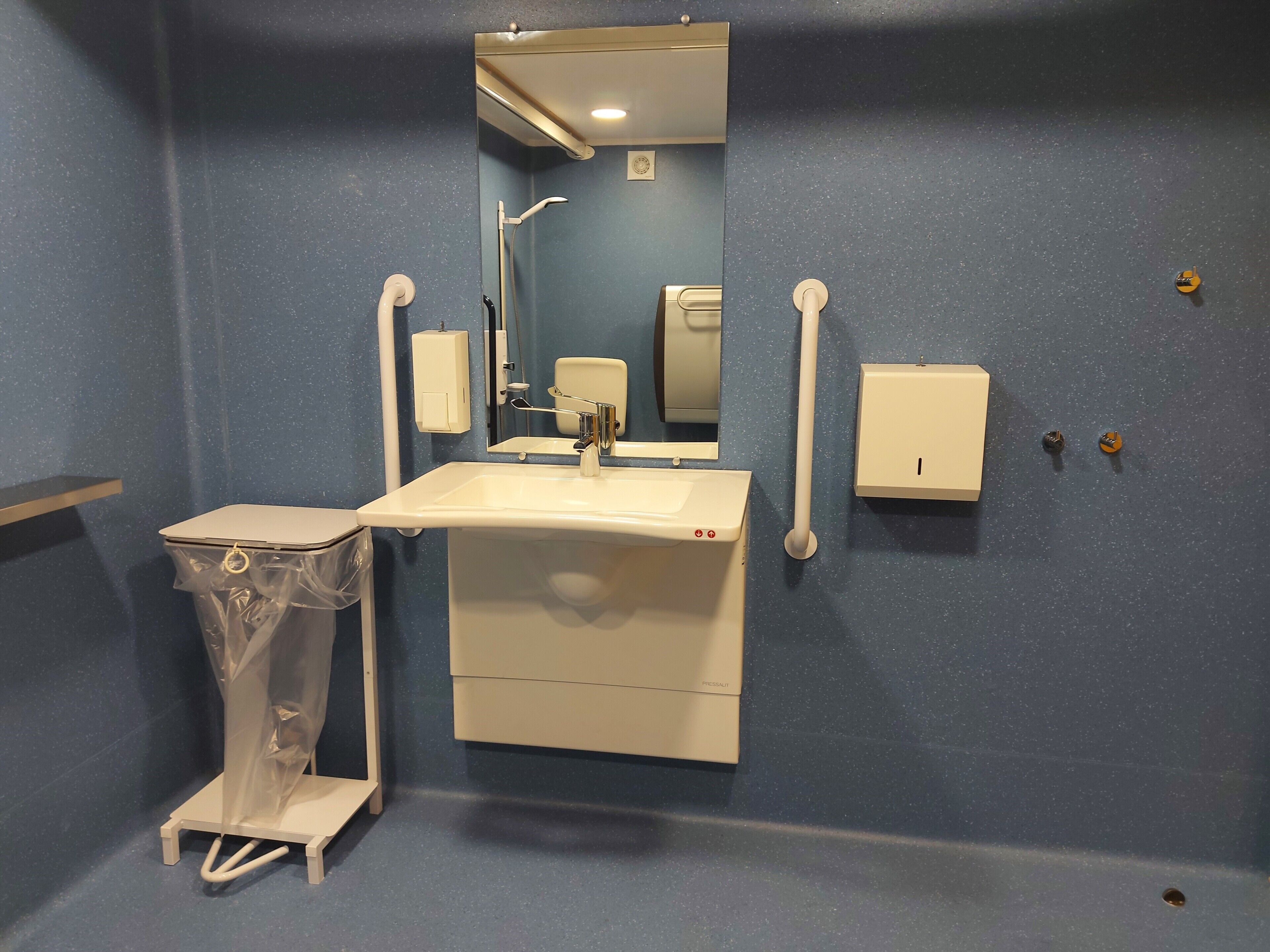 Un baño con lavamanos, espejo, secador de manos y un cubo de basura.