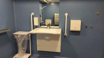 Un baño con lavamanos, espejo, secador de manos y un cubo de basura.