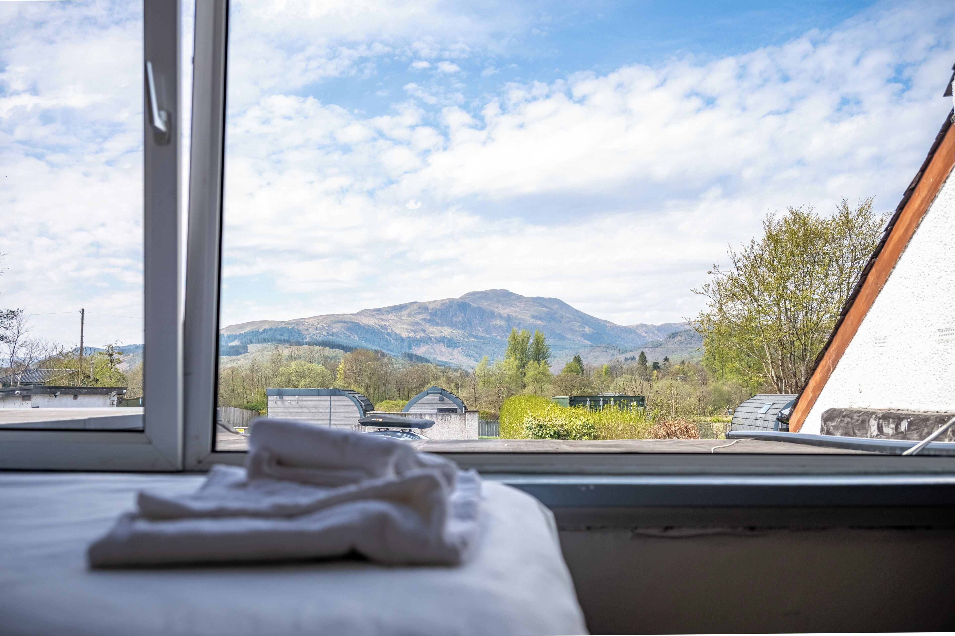 Quarto com vista para montanhas pela janela, cama com cobertor branco e teto de madeira.