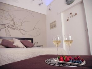 Un dormitorio con cama, un muro con un dibujo de un árbol, dos copas de vino blanco y un plato de fresas y arándanos.