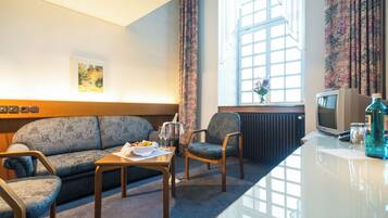 Een hotelkamer met een bank, fauteuil, een klein tafeltje met een fles en glazen, een televisie en een raam met gordijnen.