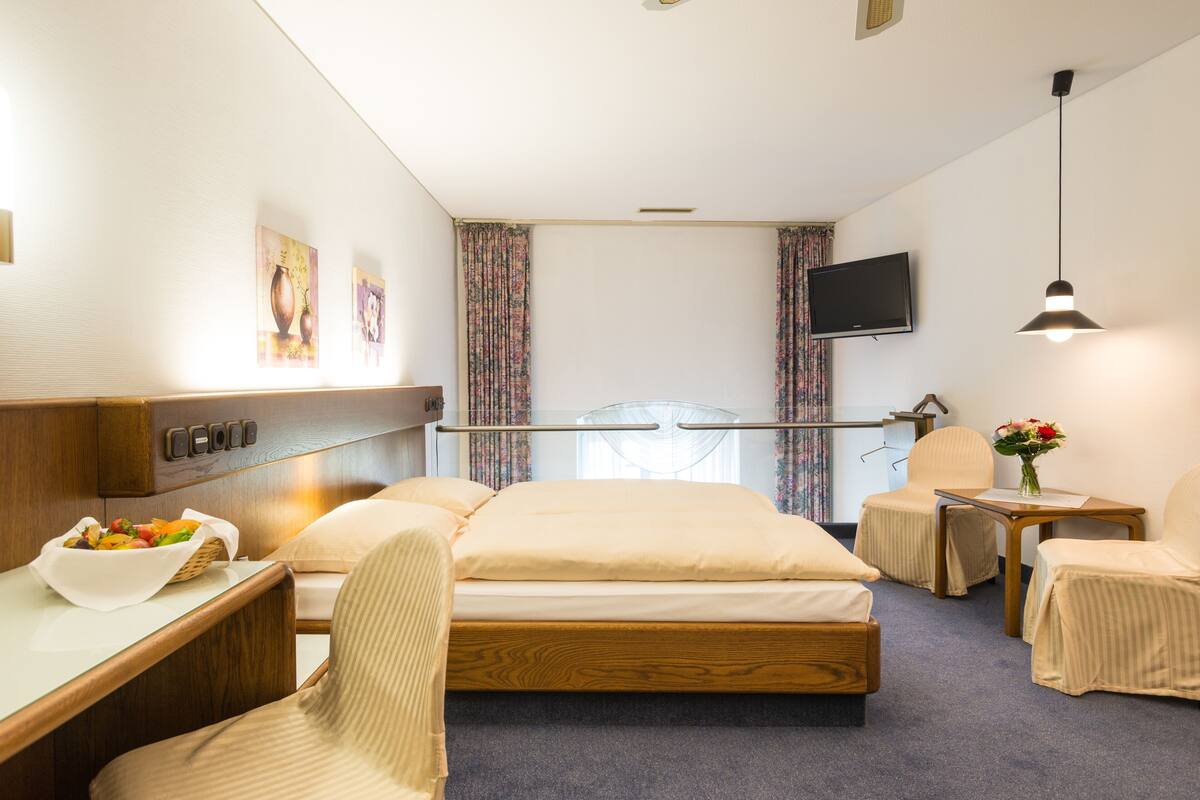 A hotel room with a bed, a desk with a bowl of fruit, a chair, a small table, a TV, and a window with curtains.