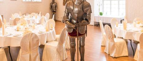 A banquet hall with round tables set for a formal event, a knight's statue in the center, and a large window providing natural light.