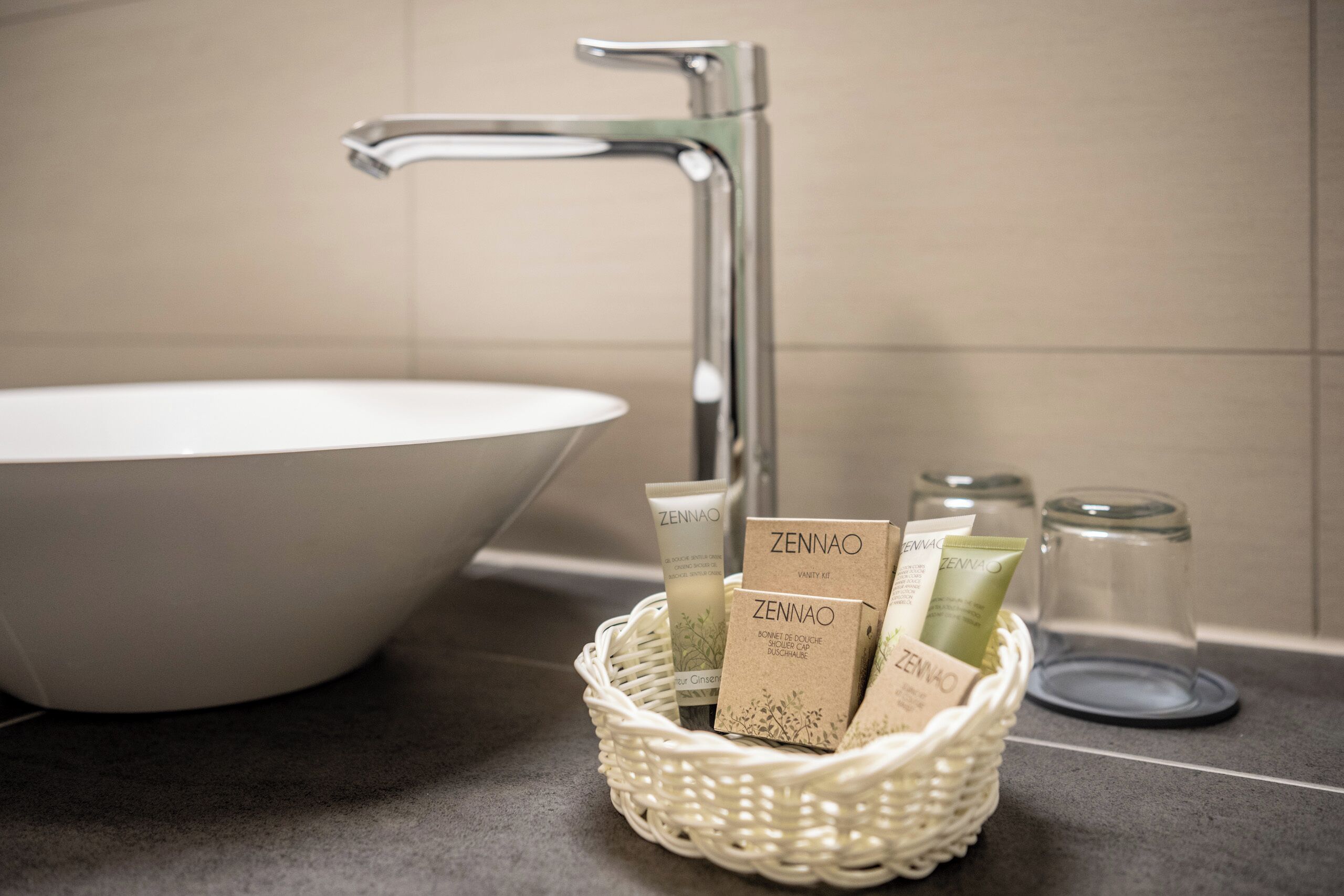 Un baño con un lavamanos blanco, un grifo cromado y una cesta de mimbre con productos para el cuidado de la piel Zennao.