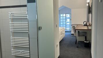 Un pasillo con una puerta blanca, un radiador blanco, una alfombra gris, una mesa de madera, un banco gris y una ventana con vistas a edificios.