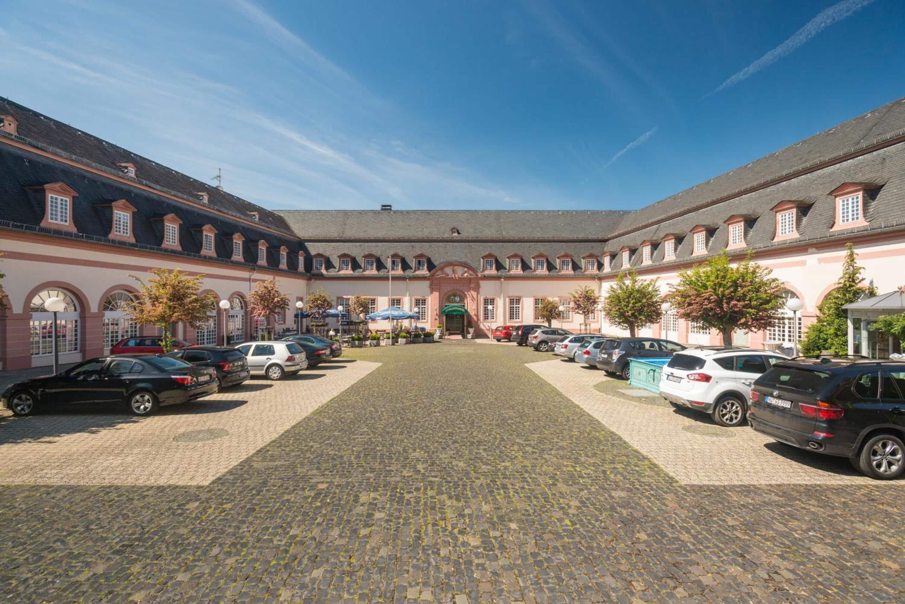 Een binnenplaats met kinderkopjes tegels, geparkeerde auto's en een gebouw met gebogen ramen en een centrale ingang.