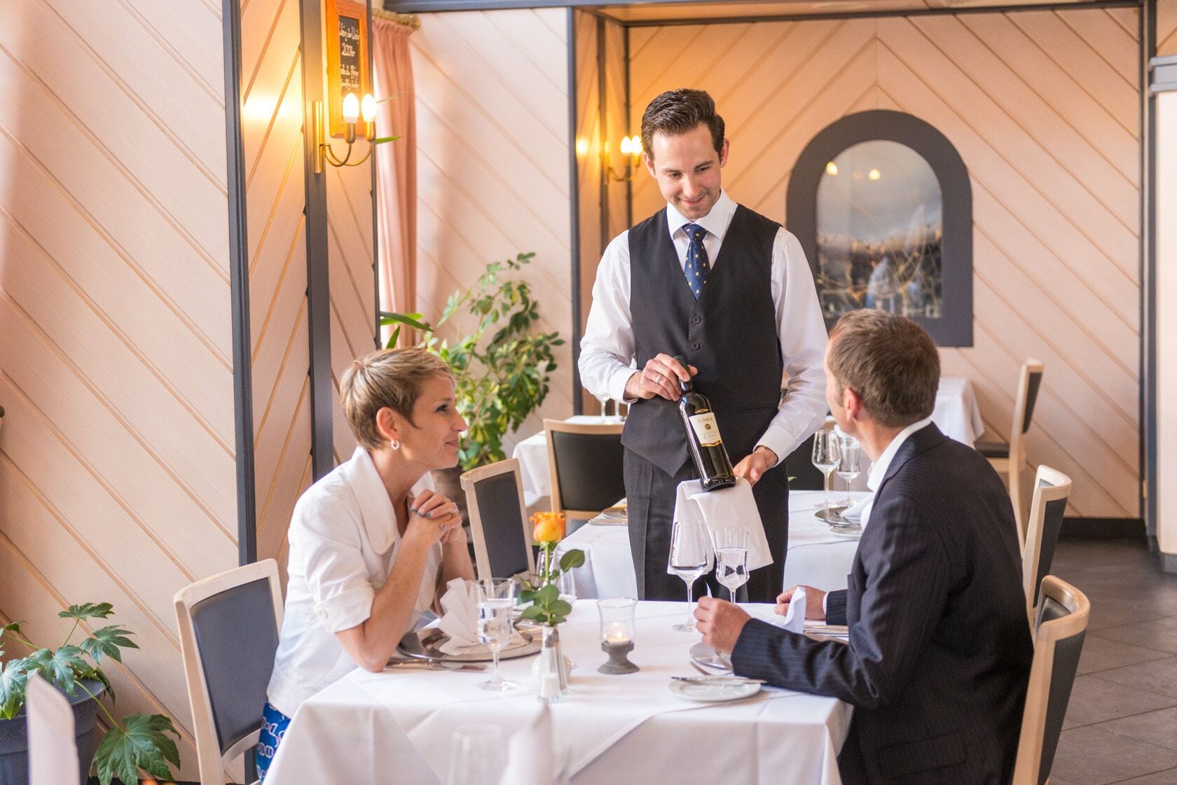 A restaurant setting with a waiter pouring wine. There are tables with white tablecloths, chairs, and a potted plant.