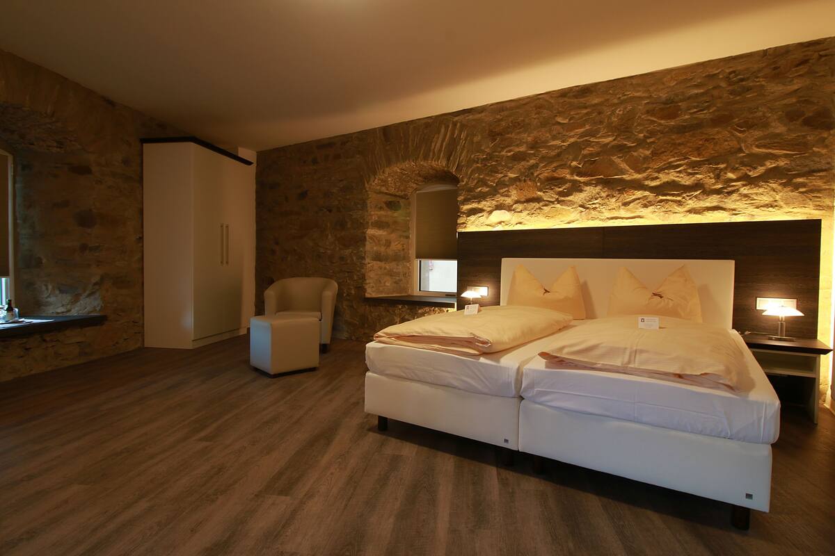 A bedroom with a stone wall, a bed, a chair, and a nightstand with a lamp.